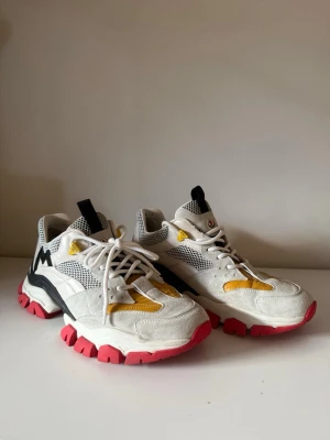 Moncler vita sneakers med färgdetaljer - Säljer ett par feta Moncler sneakers i vitt med gula och svarta detaljer samt röd sula. Skorna har mesh och mocka i ovandelen, snörning och chunky sula som sticker ut. Perfekta för dig som vill ha något unikt och sportigt i garderoben.