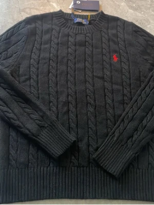 Svart stickad tröja Polo Ralph Lauren - Kabelstickad svart tröja från Polo Ralph Lauren med klassisk röd logga broderad på bröstet. Tröjan har rund halsringning. Perfekt för dig som gillar stilrena och snygga plagg.