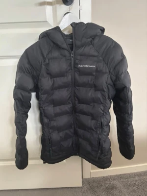 Svart dunjacka Peak Performance XS - Snygg svart dunjacka från Peak Performance med huva och dragkedja framtill. Jackan har två sidofickor med dragkedja och diskret logga på bröstet. Perfekt för kalla dagar och riktigt lätt att bära. Klassisk passform och quiltad design.
