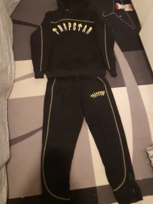 Svart Trapstar set med gula detaljer - Säljer ett svart set från Trapstar med matchande hoodie och byxor. Setet har gula och vita Trapstar-loggor samt kontrastsömmar längs ärmar och ben. Perfekt för en streetwear-look. Materialet är mjukt och bekvämt, troligen bomullsblandning.
