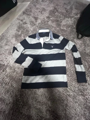 Randig långärmad pikke från Gant - Säljer en långärmad skjorta från Gant med breda marinblå och grå ränder. Skjortan har klassisk krage i kontrasterande blått tyg och Gant-logga broderad på bröstet. Tillverkad i mjuk bomull, perfekt för dig som gillar preppy stil.