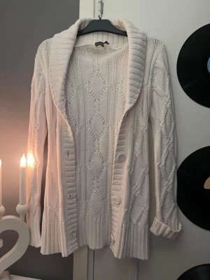 Vit kabelstickad kofta med sjalkrage - Mysig vit kabelstickad kofta med sjalkrage och knappar framtill. Koftan har ett klassiskt mönster, långa ärmar och ribbade muddar. Perfekt för lager-på-lager och chill dagar. Materialet känns mjukt och värmande.