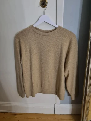 Beige stickad tröja i enkel design - En stilren beige stickad tröja med rund halsringning och långa ärmar. Tröjan har en mjuk och lite lös passform med ribbade muddar vid ärmslut och nederkant. Perfekt basplagg för lager på lager och enkel att matcha med olika outfits.