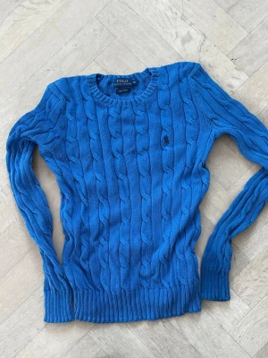 Blå kabelstickad tröja Polo Ralph Lauren - Snygg blå kabelstickad tröja från Polo Ralph Lauren med rund halsringning och långa ärmar. Tröjan har det klassiska broderade logotypen på bröstet och ribbade muddar vid ärmslut och nederkant. Perfekt för en stilren och avslappnad look.