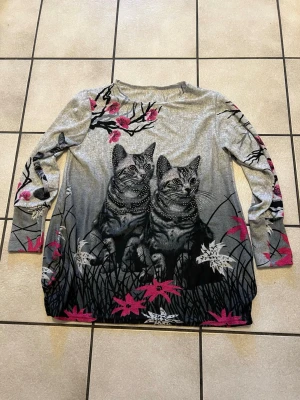 Grå långärmad topp med kattmotiv storlek L/XL - Unik grå långärmad topp med stort tryck av två katter framtill, dekorerad med blommor i rosa och vitt samt grenar. Toppen har rund hals och är tillverkad i mjukt material, perfekt för dig som gillar djur och vill sticka ut med ett coolt motiv.