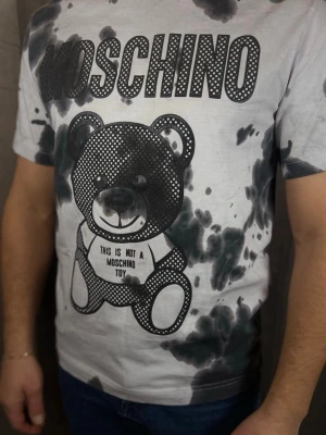 Vit Moschino t-shirt med björntryck - Säljer en vit t-shirt från Moschino med stort svart björntryck och texten 'This is not a Moschino Toy' på framsidan. T-shirten har svarta batikmönster och korta ärmar. Perfekt för dig som gillar statement-plagg och cool design.