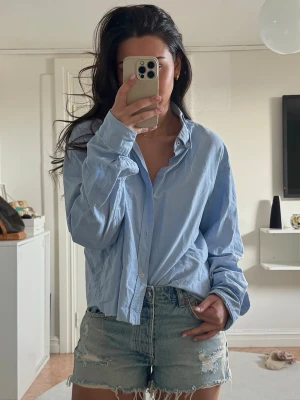 Ljusblå skjorta i bomull - Snygg ljusblå skjorta i bomull med klassisk krage och knappar framtill. Skjortan har en avslappnad passform och långa ärmar, perfekt att styla till jeans eller shorts för en chill look.