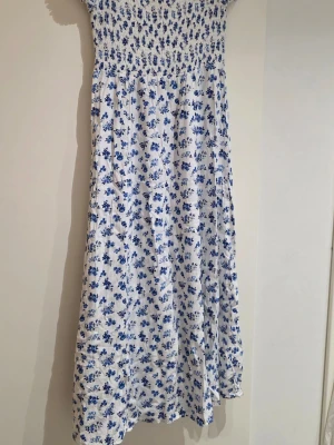Vit långklänning med blå blommor - Söt långklänning i vitt med blått blommönster. Klänningen har smock upptill och är ärmlös, vilket ger en avslappnad och somrig känsla. Perfekt för varma dagar och har en luftig passform. Materialet känns som bomull eller linne.