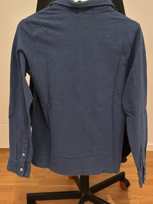 Blå långärmad polotopp från Polo Ralph Lauren - Klassisk blå långärmad polotopp från Polo Ralph Lauren med krage och knappar framtill. Tillverkad i mjuk bomull och har en diskret broderad logga på bröstet. Perfekt för en avslappnad men ändå stilren look.