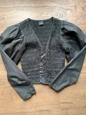 Svart croppad topp med puffärm - Trendig svart croppad topp från Gina Tricot med smockad kropp och långa puffärmar. Djup v-ringning framtill och figurnära passform i midjan. 