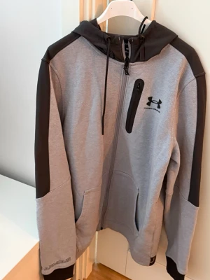 Grå hoodie från Under Armour - Snygg grå hoodie från Under Armour med svarta detaljer på axlar, huva och ärmar. Dragkedja framtill, bröstficka med zip och två sidofickor. Tillverkad i mjukt material som känns skönt mot huden. Perfekt för en sportig och avslappnad stil.