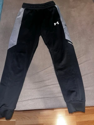 Svarta mjukisbyxor och svart hoodie från Under Armour - Svarta mjukisbyxor med gråa paneler längs sidorna och resår i midjan. Byxorna har Under Armour-logga på låret och smal passform med muddar vid bensluten. Perfekta för chill eller träning.Mjukis byxor comer med combo av en svart Under Armour hoodie. Byxorna är väldigt bra och stretchiga, på byxorna gick tyget lite sönder men jag sydde och man ser verkligen inte ens.