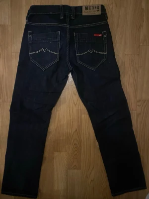 Mörkblå jeans från Mustang - Snygga mörkblå jeans från Mustang med raka ben och kontrastsömmar. Klassisk femficksmodell med detaljerade bakfickor och läderpatch i midjan. Perfekta för en avslappnad och stilren look. Tillverkade i slitstark denim.