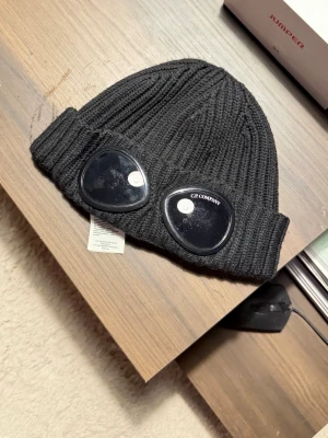 Svart mössa med goggles från C.P. Company - Säljer en svart ribbstickad mössa från C.P. Company med ikoniska goggles framtill. Mössan är i mjukt material och har en uppvikt kant. Perfekt för dig som vill sticka ut med en streetwear-inspirerad stil.