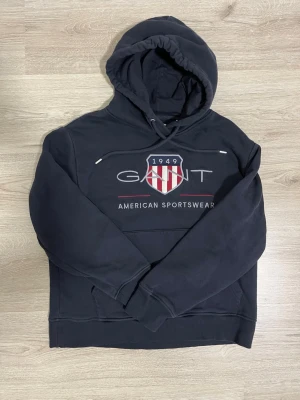 Svart hoodie från GANT med tryck - Mörkblå hoodie från GANT med huva och dragsko. På bröstet finns ett stort tryck med GANT-logga, amerikansk sköld och texten '1949 American Sportswear'. Tillverkad i mjukt material, perfekt för en avslappnad stil.