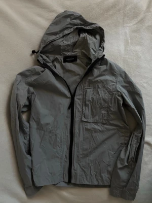 Ljusgrå Peak Performance vindjacka  - Ljusgrå vindjacka från Peak performance med huva, svart dragkedja och bröstficka. Jackan är tillverkad i ett lätt material som passar perfekt för blåsiga dagar.