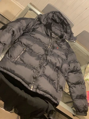 Svart pufferjacka med huva - Säljer en svart pufferjacka med huva och dragkedjor framtill samt på fickorna. Jackan har en quiltad design och en liten röd logga på bröstet. Perfekt för kalla dagar och riktigt skön att bära. Materialet är glansigt och ger en modern look.