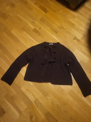 Vinröd kofta med rosettknäppning - Mysig vinröd kofta med två dekorativa rosetter framtill som knäppning. Koftan har lång ärm och är stickad i ett mjukt material, perfekt för lager-på-lager. Enkel och stilren design som passar till många outfits.