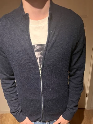 Mörkblå ziptröja med ståkrage - Mörkblå stickad ziptröja med ståkrage och hel dragkedja framtill. Tröjan har lång ärm och ribbade muddar vid ärmslut och nederkant. Perfekt för lager-på-lager och enkel att matcha med jeans eller t-shirt.