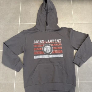 Grå Saint Laurent hoodie  - Snygg grå hoodie från Saint Laurent med huva och känguruficka. Framsidan har ett stort tryck med texten 'SAINT LAURENT' och röda samt vita ränder, samt ett emblem i mitten. Perfekt för en avslappnad och trendig stil.