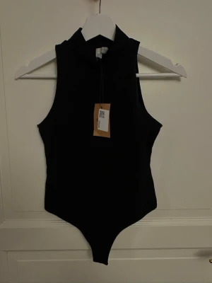 Svart ärmlös body med hög krage - Stilren svart body med hög krage och ärmlös design. Perfekt att styla till olika outfits. Mjukt och stretchigt material för en bekväm passform. Enkel och modern look.