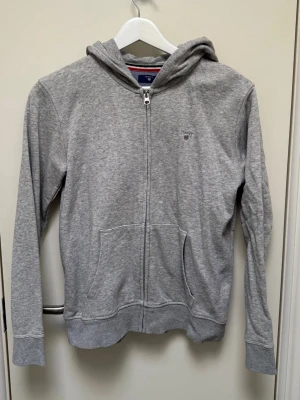 Grå zip hoodie från Gant - Nypris: ca 1000 kr knappt använd! snyggt grå hoodie från Gant med dragkedja framtill och klassisk huva. Tröjan har två stora fickor på magen och en liten Gant-logga broderad på bröstet. Tillverkad i mjukt bomullsmaterial och har ribbade muddar vid ärmslut och nederkant.