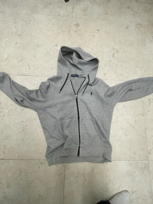 Grå zip-hoodie från Polo Ralph Lauren - Snygg grå hoodie med dragkedja från Polo Ralph Lauren. Klassisk design med huva, svarta snören och två fickor framtill. Liten broderad logga på bröstet. Perfekt för en avslappnad och stilren look.