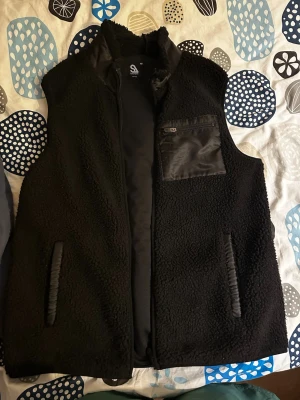 Svart fleeceväst från lager 157  - Säljer en svart fleeceväst från Salomon med mjukt teddy-material, dragkedja framtill och detaljer i nylon på fickor och bröstficka. Perfekt lager för kyliga dagar och enkel att matcha med olika outfits.