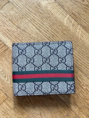 Gucci GG Supreme plånbok - Snygg plånbok från Gucci i GG Supreme-mönster med klassiska gröna och röda ränder. Plånboken är i grått och svart med detaljer i tyg och läder. Perfekt för dig som gillar lyxiga accessoarer med ikonisk design.