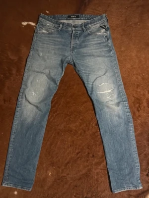 Blå Jeans från Replay 31/32 - Säljer ett par blå jeans från Replay med slitningar på knäna och snygg tvätt. Klassisk femficksmodell med raka ben och detaljerade sömmar på bakfickan. Perfekta för en avslappnad och trendig look. Storlek 31/32. Snabb Leverans!