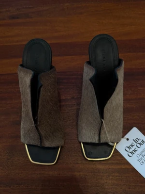 Céline klackar  - Unika sandaler från Celine med öppen tå och fyrkantig sula. Ovandelen är klädd i brunt ponnyhår och har en cool, asymmetrisk design. Svart innersula och gulddetaljer runt sulkanten ger en lyxig känsla. Perfekta för dig som vill sticka ut.