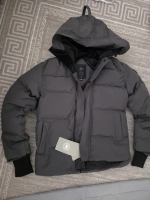 Mörkgrå pufferjacka från Canada Goose - Säljer en mörkgrå pufferjacka från Canada Goose i storlek L. Jackan har huva, stora fickor och grov dragkedja med knappar. Materialet är  dun, perfekt för kalla dagar. Stilen är clean och modern med ribbade muddar vid ärmslut.