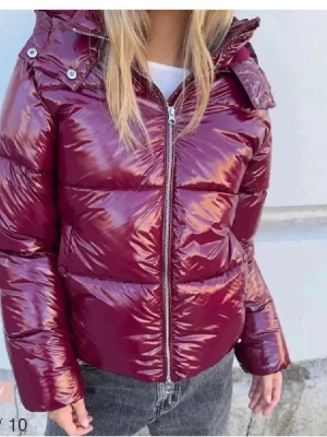 Nelly jacka - Säljer en vinröd pufferjacka med glansig finish och dragkedja framtill. Jackan har hög krage, avtagbar huva med tryckknappar och breda kanaler. Perfekt för kalla dagar och riktigt snygg till jeans.