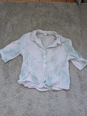 Ljusgrön och vit skjorta med knyt - Säljer en luftig skjorta i ljusgrönt och vitt med knyt framtill. Skjortan har korta ärmar, krage och knappar hela vägen. Perfekt för varma dagar och har en avslappnad, trendig look.