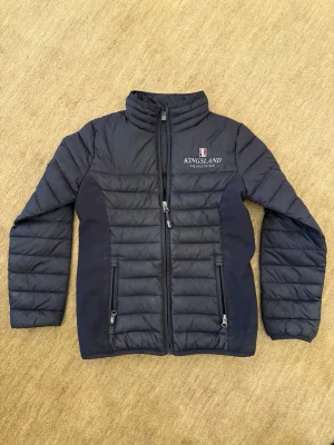 Mörkblå dunjacka från Kingsland - Snygg mörkblå dunjacka från Kingsland Equestrian med quiltade paneler och hög krage. Jackan har dragkedja framtill, två fickor med dragkedja och diskret logga på bröstet. Perfekt för kalla vinterdagar och har en sportig look. Storlek 134/140