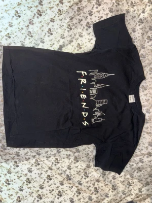 Svart Friends t-shirt med tryck - Cool svart t-shirt med Friends-tryck och stadssilhuett på bröstet. Klassisk passform med rund hals och korta ärmar. STORLEKEN ÄR 12Y