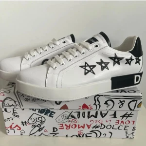 Dolce & Gabbana vita sneakers med stjärnor - Snygga vita sneakers från Dolce & Gabbana med svarta stjärnor och grafiska detaljer på sidorna. Skorna har en chunky sula, svarta och vita kontraster samt logga på hälen. Tillverkade i slätt läder med vita skosnören och cool streetstyle-vibe.
