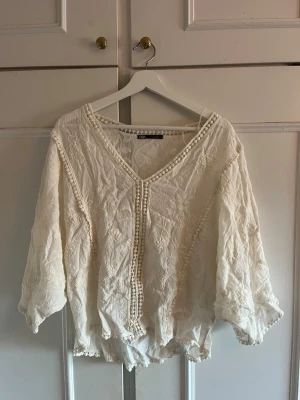 Vit boho-blus från Zara - Superfin vit blus från Zara med v-ringning och broderade detaljer. Blusen har trekvartsärmar, lös passform och dekorativa bollar längs kanterna. Perfekt för en avslappnad och trendig look.