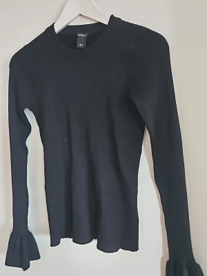 Svart stickad tröja med volangärm - Snygg svart stickad tröja från Lindex med långa ärmar och trendiga volangdetaljer vid ärmsluten. Tröjan har en rund halsringning och är i ett mjukt, stretchigt material som sitter skönt på kroppen. Perfekt för dig som gillar stilrena plagg med en twist.