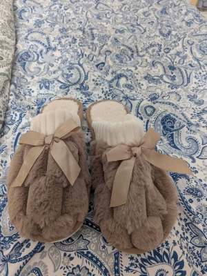 Beige fluffiga tofflor med rosett - Supersöta beige tofflor med fluffig fuskpäls, vita stickade muddar och stora satinrosetter framtill. Perfekta för att mysa runt hemma och hålla fötterna varma. Tofflorna har öppen häl och mjuk innersula för extra komfort.