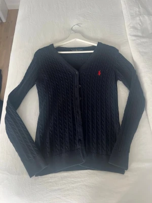 Mörkblå kabelstickad kofta Ralph Lauren - Mörkblå kabelstickad kofta från Ralph Lauren med V-ringning och knäppning framtill. Klassisk design med röd broderad logga på bröstet. Långa ärmar och ribbade muddar. Perfekt för lager-på-lager och snygg till jeans.