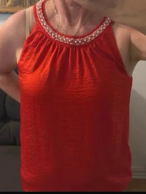 Röd ärmlös topp med pärldetaljer - Snygg röd ärmlös topp med rund halsringning dekorerad med pärlor och broderade detaljer. Toppen har en lätt skrynklig struktur och knäpps i nacken. Perfekt för dig som vill sticka ut med färg och detaljer. Passar S/M.