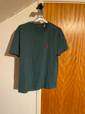 Mörkgrön t-shirt från Polo Ralph Lauren - Klassisk mörkgrön t-shirt från Polo Ralph Lauren med röd broderad logga på bröstet. T-shirten har rund halsringning och korta ärmar. Tillverkad i mjuk bomull för en skön känsla och enkel stil. Kan tänka mig att gå ner i pris.