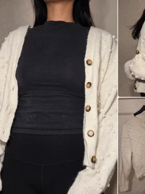 Vit kofta med träknappar KappAhl - Säljer en krämvit kofta från KappAhl med träknappar och små bollar i stickningen. Koftan har v-ringning, ribbade muddar och en avslappnad passform. Perfekt att slänga över en t-shirt för en chill look.