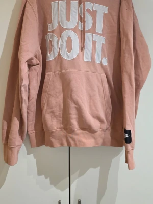 Nike rosa hoodie med tryck - Säljer en snygg rosa hoodie från Nike med stort vitt 'JUST DO IT.'-tryck på bröstet och en svart Nike-logga på ärmen. Hoodien har huva med dragsko och en stor magficka. Perfekt för dig som gillar streetwear och sportig stil.