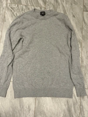 Grå basic tröja från H&M - En enkel och stilren grå tröja från H&M med rund halsringning och långa ärmar. Tröjan har ribbade muddar vid ärmslut och nederkant, perfekt för en clean och avslappnad look. Tillverkad i mjukt material för extra komfort.