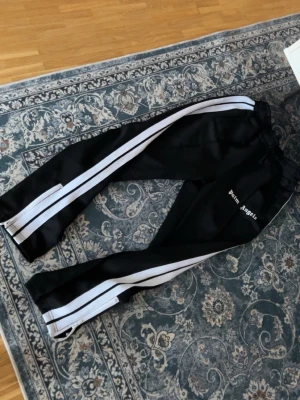 Palm Angels svarta mjukisbyxor - Svarta mjukisbyxor från Palm Angels med vita sidoränder och logga tryckt på låret. Byxorna har en avslappnad passform och resår i midjan. Perfekta för en streetwear-look och tillverkade i ett glansigt, sportigt material.