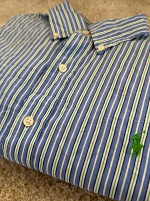 Blåvit randig skjorta från Polo Ralph Lauren - Klassisk skjorta från Polo Ralph Lauren i blått med vita och gröna ränder. Skjortan har knappar framtill och den ikoniska gröna broderade loggan på bröstet. Perfekt för en stilren look och tillverkad i mjuk bomull.