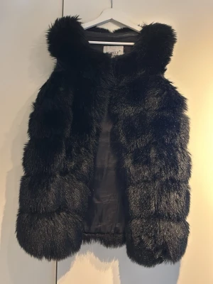 Svart fluffig pälsväst från Lovely st M - Snygg svart pälsväst från Lovely med glansig finish och breda, fluffiga paneler. Västen är ärmlös och har en mjuk insida i tyg. Perfekt för att lyfta din outfit med en lyxig touch, finns även fickor 