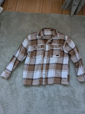 Beige och vit rutig overshirt - Overshirt i beige och vitt rutigt mönster med bruna detaljer. Två bröstfickor med lock och knappar framtill. Klassisk krage och långa ärmar med knapp vid ärmslut. Tjockare material som ger en avslappnad och trendig look.